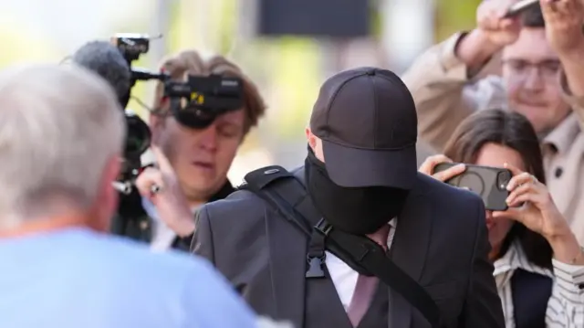 Robert Bush points his head down while surrounded by press. He is wearing a dark suit, baseball cap and face covering.