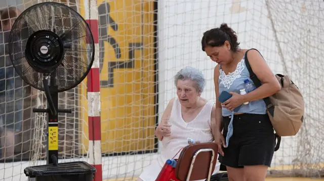An old woman sits in a chair next to a younger woman. They are in a sports centre in front of a goal, with a large fan in the foreground