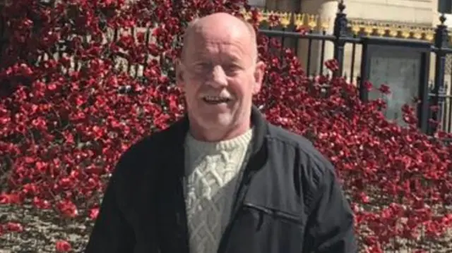 Man in black jacket and cream jumper in front of a red bush