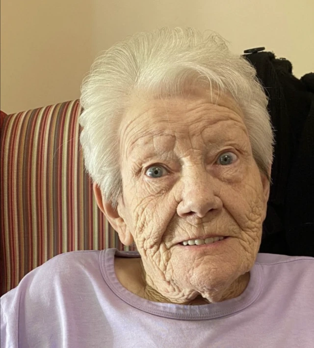 An elderly woman sat in front of striped wallpaper