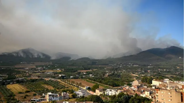A town in the foreground in sunshine, with a large cloud of smoke rising from the green hills beyond