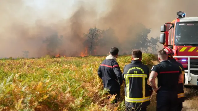 Firefighters are shown standing in front of scrub beside a fire truck, as flames and smoke billow among trees in the distance.