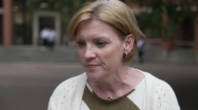 A woman with a blonde bob is looking down to the side and wearing a white cardigan, green and white top and silver necklace. The background is blurred.