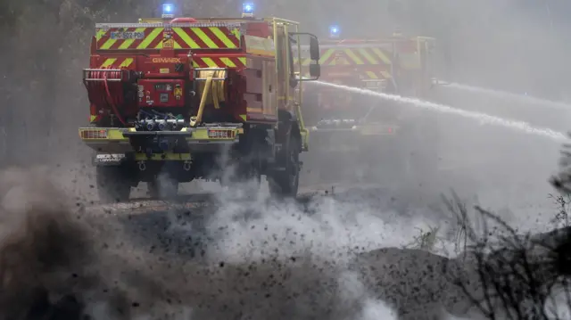 Two fire engines travel down dirt road and spray water into scrubland