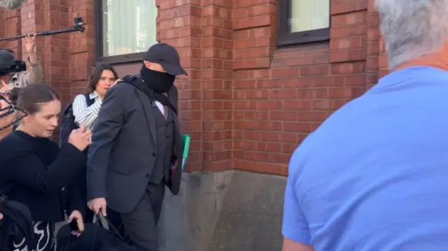 Robert Bush walks outside a court building wearing a dark suit, baseball cap and face covering. Members of the press can be seen around him.