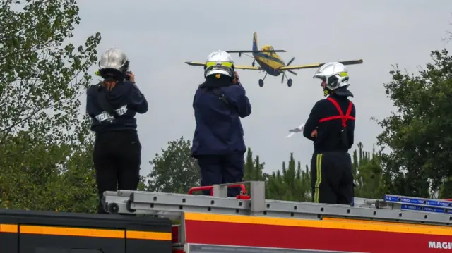 People wearing helmets standing on top of a fire engine as a small plane flies overhead
