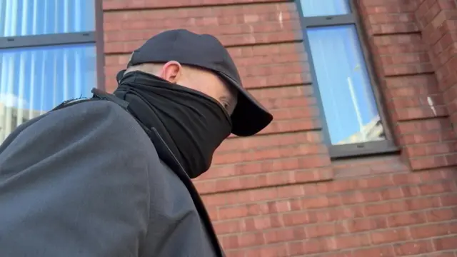 Robert Bush walks outside court wearing a baseball cap and face covering. A building with a red brick wall is visible in the background.