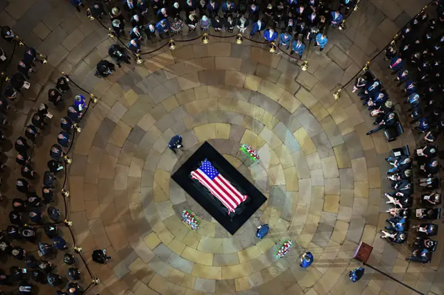 Casket with US flag draped over it on black platform, yellowed stone flooring, circle of people sitting