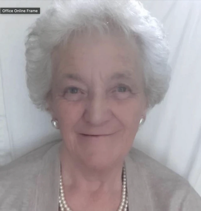 A woman smiles at the camera she is wearing a pearl necklace and earrings and has grey short hair