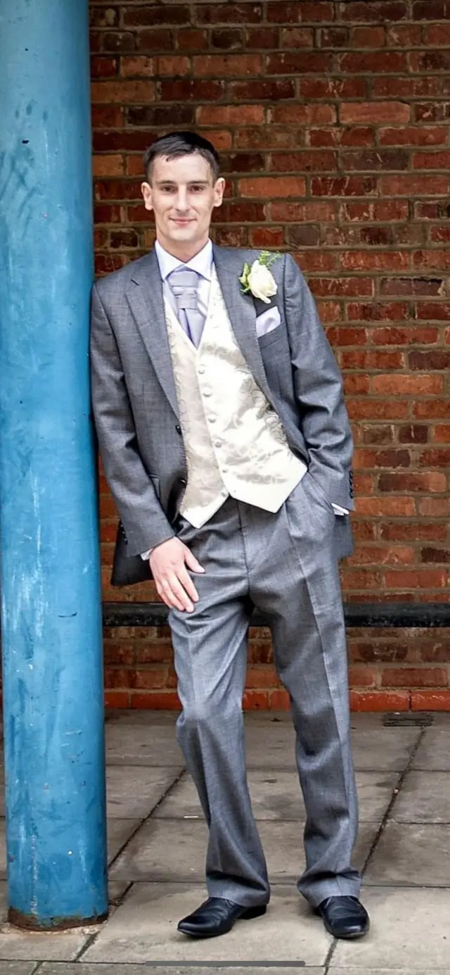 A young man is wearing a formal grey suit with a cream waistcoat and a flower in his buttonhole. He is leaning against a blue coloured column in front of a red brick wall. he is smiling and has short, neat, dark hair