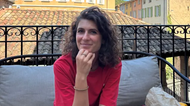A woman with brunette curly hair wearing a red top, smiling. She's sitting on what appears to be a balcony, on an iron outdoors couch with grey cushions