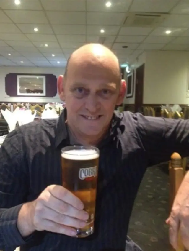 A bald man wearing a black shirt is holding a pint of beer and smiling at the camera, he is sitting in a restaurant and tables and chairs are visible behind him