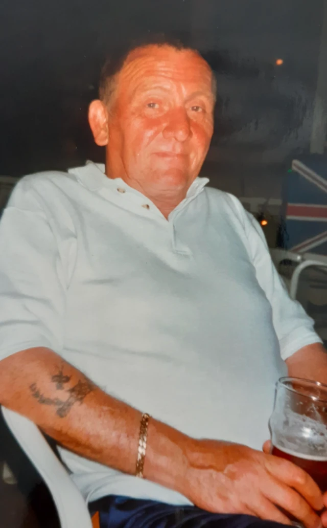 A man sits with a pint of beer and smiles to the camera, he is wearing a pale polo shirt and is tanned with tattoos on his forearm and a gold chain on his wrist