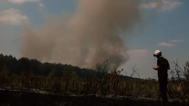 Smoke rises above pine trees in Saint-Jean-d'Illac. An emergency worker in a white helmet checks his phone nearby