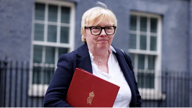 Angela Eagle outside 10 Downing Street. She has short blonde hair and is wearing black-framed glasses, a blue blazer and a white shirt. She is holding a red folder.