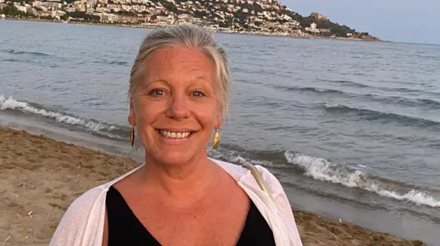 A woman smiling, with blonde hair, gold earrings and a sea behind her