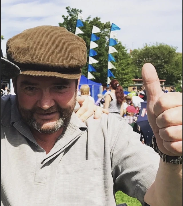 A man with a salt and pepper grey and brown beard is wearing a brown corduroy cap and giving the camera a thumbs-up gesture