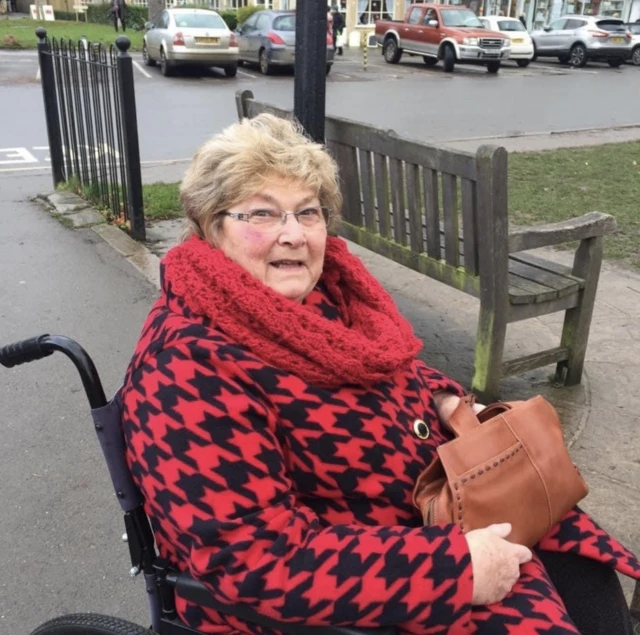 a woman in a wheelchair, she has short blonde hair and is wearing a red and black check top coat and a red scarf