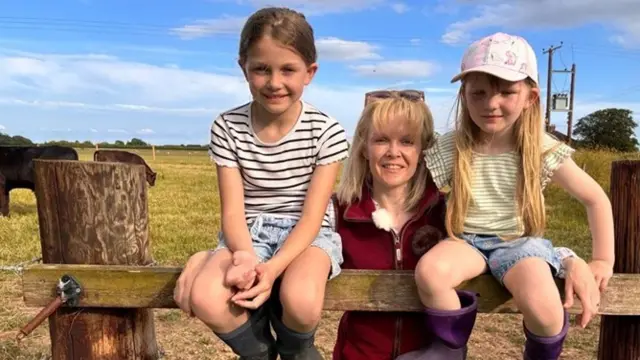 Heather Oldfield stands in the centre of the image wearing a burgundy fleece gilet and sunglasses on her head. On either side of her, sitting on a wooden fence is a young girl. They are in a field with cattle behind them.