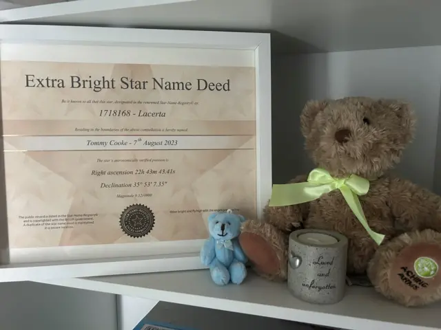 a framed certificate showing a star named after Tommy Cooke, alongside it are a tiny blue teddy and a tealight candle and a larger teddy with a green bow around its neck