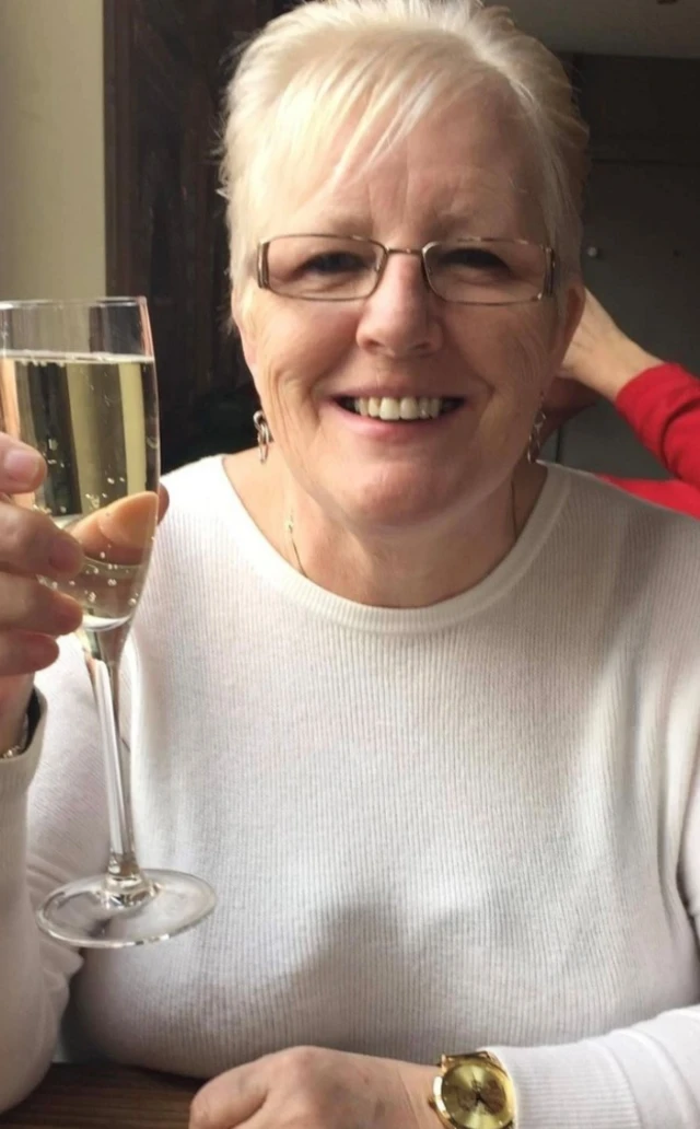 A woman with blonde hair is smiling at the camera while holding a wine glass, she is wearing glasses and has gold earrings and a necklace on.