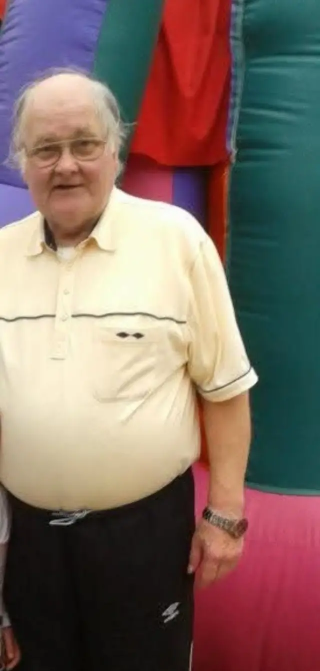 A man in a yellow polo shirt and black tracksuit bottoms is wearing oval-rimmed glasses and has short grey hair. He is stood up.