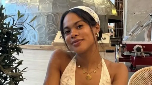 A young woman smiling, with a gold necklace on and a white flowery top