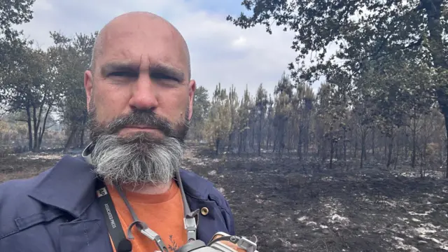 Mark Carlson stands in the left of the frame. He is wearing a navy jacket and there is a burned landscape behind him.