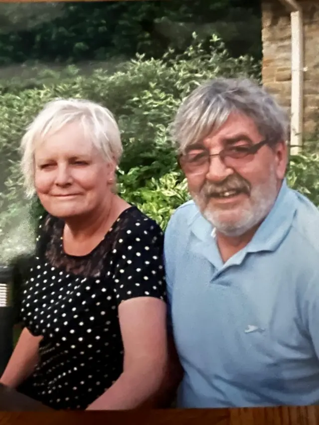 A woman in a black top with white dots has short white hair and is sat next to a man in a blue polo shirt with grey hair and glasses.