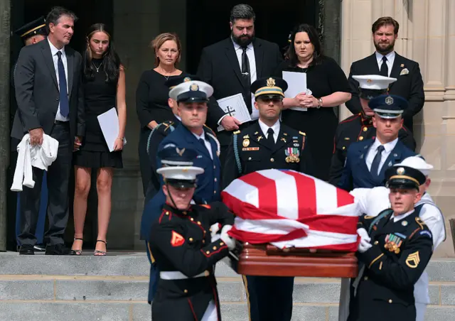 A casket draped in a US flag is carried down the steps of a cathedral by military personnel as the family of the deceased watch on
