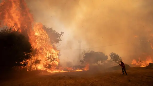 A large flame and orange sky can be seen as a firefighter sprays water.