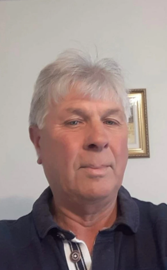 A portrait of a man with grey hair, he is wearing a blue polo shirt