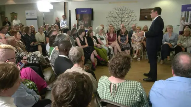 Andy Burnham, who has short dark hair and glasses, speaks while wearing a business suit to a group of people seated around him