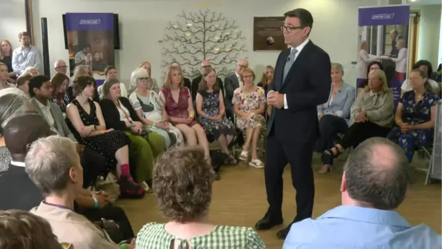 Andy Burnham, who has short dark hair and glasses, speaks while wearing a business suit to a group of people seated in a circle around him