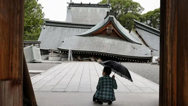 A person holding an umbrella crouches down in front of the collapsed worship hall of Yatshushiro Shrine. The ceiling of the shrine is on the floor and its buildings are now at a tilt