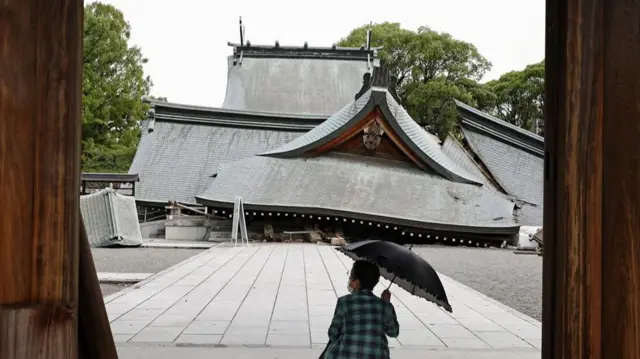 A woman with an umbrella squats in front of a damaged traditional building