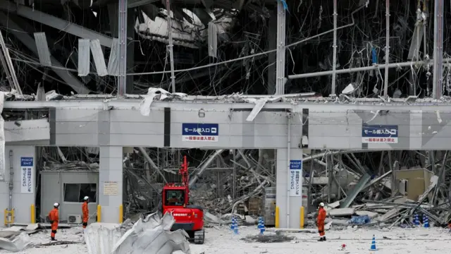 A red digger and several emergency workers can be seen beneath the exploded front of the shopping mall. Wires and pipes can be seen dangling from the first floor while large bits of concrete debris can be seen at the bottom of the building
