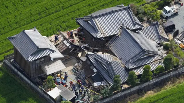 Several collapsed buildings, wooden fences and tiled roofs can be seen from above. Around a dozen emergency workers can be seen searching the rubble
