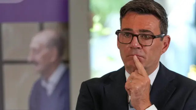 Andy Burnham, who has short dark hair and glasses, holds his index finger to his chin in a contemplative pose
