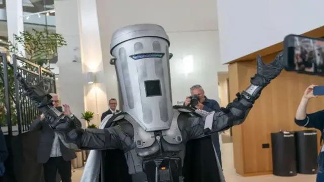 Person wearing a grey costume with a cape and what looks like body armour and a bin over their head