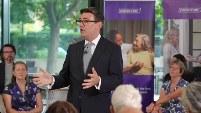 Andy Burnham, who has short dark hair and glasses, speaks while wearing a business suit, with people seatedbehind him listening