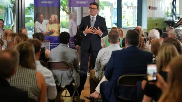 Andy Burnham, who has short dark hair and glasses, speaks while wearing a business suit