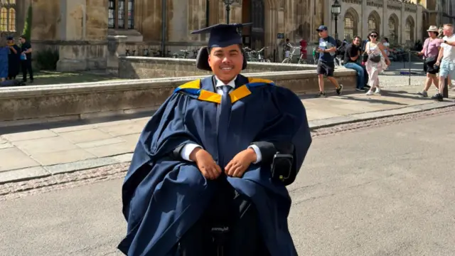 A man in a navy graduation gown sits in a mobility chair in front of an old building. He is grinning.