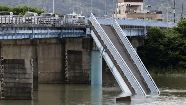 One side of a bridge lies in the river beneath it. A road bridge remains intact next to it.