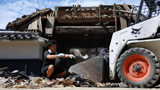 A person removes debris from a collapsed gate at a temple. He can be seen throwing a piece of debris into the front of a forklift truck