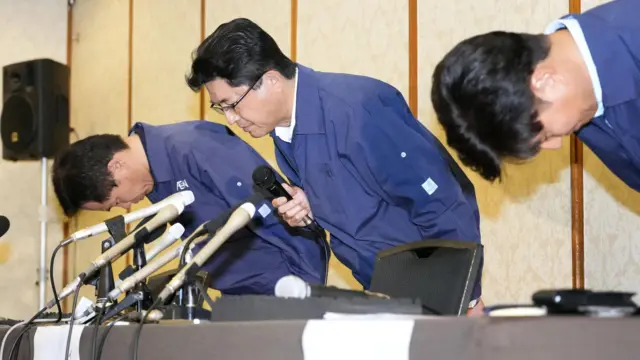 Akio Yoshida bows after a press conference. He wears a navy jacket and glasses and is holding a microphone