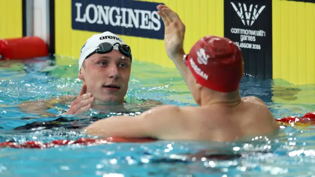 Pieter Coetze, wearing a white swimming cap with his goggles on his forehead, shakes hands with England's Oliver Morgan, who is wearing a red cap and has his back to the camera, after the Commonwealth Games semi-final