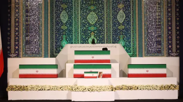 Five wooden coffins painted in the colours of the Iranian flag rest on top of a white platform in Tehran. At the very top of the platform, is Ayatollah Khamenei's coffin with a black turban resting in the right hand corner. Below it, a matching coffin and a smaller one, a picture of a young girl in pink clothing in a white frame propped against it