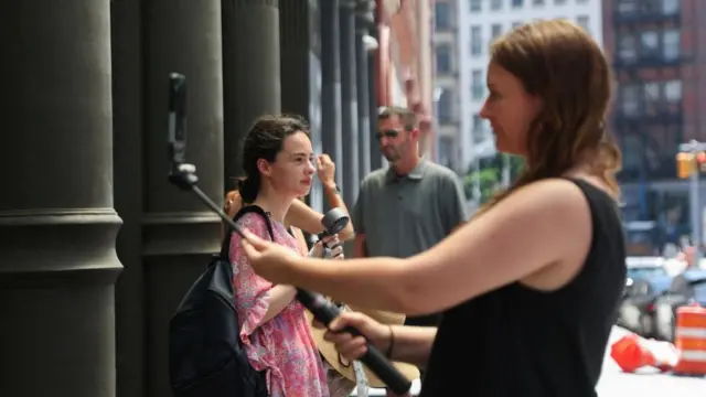A woman takes a selfie using a selfie stick outside Manhattan. A second woman uses a fan to cool off in the background