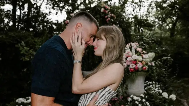 Taylor Swift places both her hands on Travis Kelce' face surrounding by flowers. An engagement ring can clearly be seen on her finger